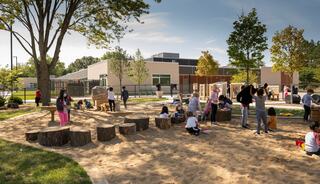 Children play in a sandy area surrounded by trees and logs, with a modern building in the background under a clear blue sky.