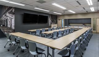 A modern conference room features a long table surrounded by chairs, dual screens, and a stylish wall mural, with a kitchenette area.