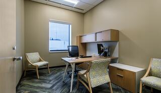 A simple office space featuring a desk, ergonomic chair, two patterned chairs, and a window allowing natural light.