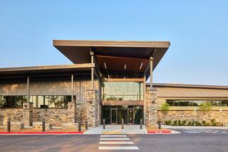 A modern building features a striking entrance with a large overhang, stone accents, and well-manicured landscaping under a clear blue sky.