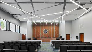 A modern courtroom featuring sleek design, rows of chairs, ample natural light, and a prominent seal on the wooden panel.