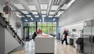 A modern, open kitchen area features a central island, seating, and large windows, with people interacting and engaging in various activities.