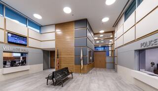 A modern municipal lobby features seating, wall panels, and signage for Village Hall and Police, with large windows allowing natural light.