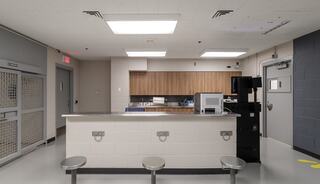 A minimalistic, institutional space featuring a service counter, metal seating, and a kitchenette area with basic appliances.