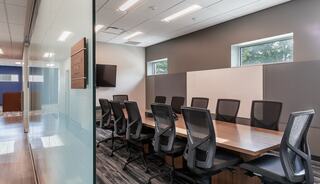 A modern conference room featuring a long table, ergonomic chairs, windows, and a wall-mounted TV for presentations.