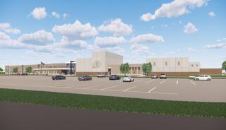A modern school building with a spacious parking lot, surrounded by greenery under a clear blue sky, featuring clouds in the background.