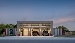A modern fire station with two fire trucks visible in large bays, featuring a sleek design and a welcoming, functional facade.