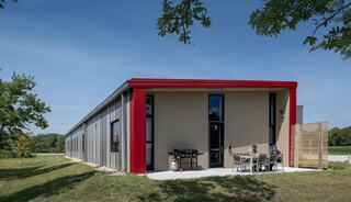 A modern fire station with a red and gray exterior, featuring patio seating and lush greenery surrounding it under a clear blue sky.