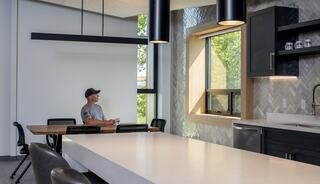 A man sits at a table in a modern kitchen, featuring large windows, sleek cabinetry, and stylish lighting, creating a bright ambiance.