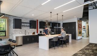 A modern kitchen features sleek black cabinetry, a large island, and two people engaged in conversation, surrounded by stylish decor.