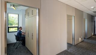 A man sits at a desk in a small office space, surrounded by light-colored walls and a window that overlooks greenery.
