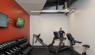A fitness space features a treadmill, weights, and a TV showing sports, with a person exercising on the treadmill.