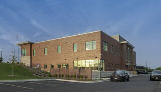 A modern two-story brick building with large windows, surrounded by landscaped green spaces and a parking lot, under a clear blue sky.