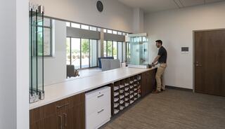 A clean, modern reception area features a man at a counter, large windows allowing natural light, and organized storage below.