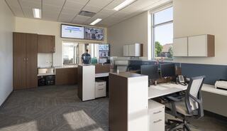 A modern office featuring a collaborative workspace with desks, monitors, and cabinets, illuminated by natural light through large windows.