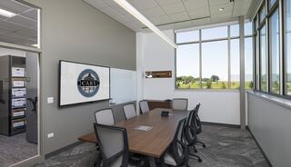 Modern conference room with large windows, a wooden table, and ergonomic chairs, featuring a wall-mounted screen and official logo.