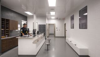 A modern, sparse reception area features a police officer at a desk, lockers, and information displays along clean, bright walls.