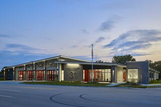 A modern fire station features a sleek design with large windows and bright red doors, set against a twilight sky.