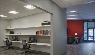 A modern office space featuring two workstations, a shelf with books, and a cozy seating area with red accents. Natural light streams in.
