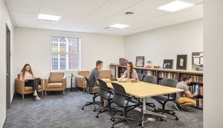A modern study space features a seating area, a collaborative table, and a person organizing books on a shelf. Natural light brightens the room.
