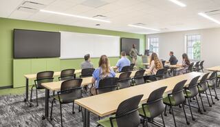 A modern classroom features a teacher at the front, with students seated at long tables, large screens, and a bright, green accent wall.