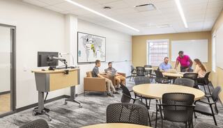 A modern classroom with round tables, a presentation station, and students collaborating. A large world map is displayed on the wall.