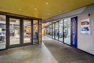 A modern campus corridor featuring glass doors leading to a store, with inviting lighting and a spacious seating area visible ahead.