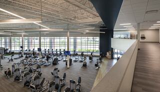 A spacious, modern gym featuring cardio machines and strength-training equipment, flooded with natural light from large windows.