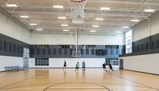 A spacious indoor gym features a basketball court with players engaged in a game, surrounded by large windows and benches.