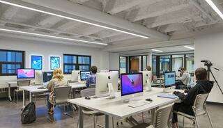 A modern computer lab features several students working on iMacs, with a neutral color palette and ample natural light from large windows.