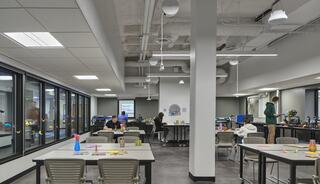 A modern classroom features students engaged in activities at tables, with large windows, bright lighting, and tech equipment in the background.