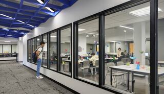 A person observes a bright, modern classroom through glass walls, where students are engaged in collaborative activities at desks.