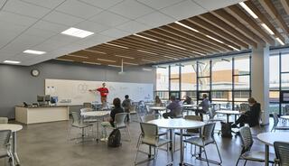 A modern classroom features a teacher presenting in front of a whiteboard, with students seated at round tables, large windows letting in natural light.