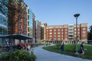 A modern campus setting features multiple buildings, pathways, greenery, and students walking or biking, creating a vibrant collegiate atmosphere.
