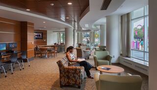 A cozy lounge features a woman reading, surrounded by modern seating and bright windows, creating a welcoming atmosphere for relaxation.