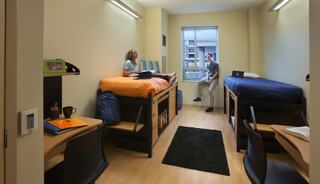 A cozy dorm room features two lofted beds, study desks, and a window view, with one student sitting and another standing by the door.