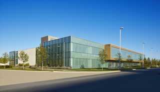 A modern building with large glass windows and a sleek design, surrounded by landscaped greenery under a clear blue sky.