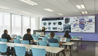 A classroom setting features a lecturer presenting an exploded view of an engine on a screen, with students engaged at their desks.