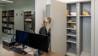 A woman inspects an open cabinet in an office space filled with shelves, boxes, and a computer setup nearby.
