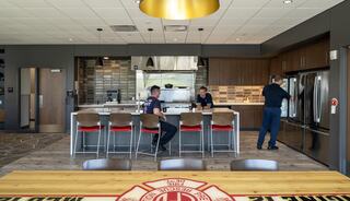 A modern kitchen with a large dining table, featuring firefighters at a bar counter and prepping food, showcasing a welcoming communal space.
