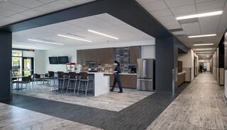 A modern kitchen area with sleek furniture, large windows, and an open layout, leading to a hallway in a stylish workspace.