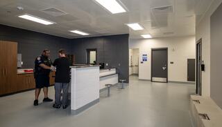 A police officer and a person interact in a clean, modern room with lockers and a reception area, featuring gray walls and fluorescent lighting.