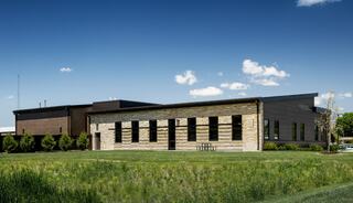 A modern building with a mix of stone and dark materials, surrounded by green grass and trees, under a clear blue sky with a few clouds.