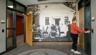 A woman in an orange shirt approaches open doors, with a historical mural of a building and people sitting in chairs on the wall behind her.