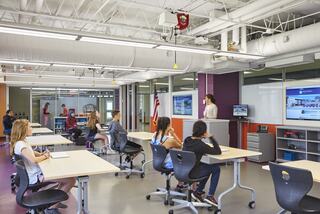 A modern classroom features students at desks facing a teacher and a screen, with bright decor and collaborative spaces in the background.