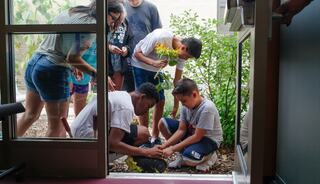 A group of kids gathers by a doorway, with two boys kneeling and examining something on the ground while others observe nearby.