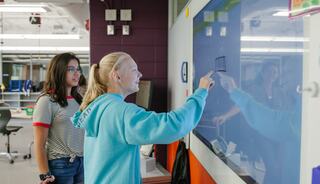 Two students engage with a digital board, one pointing while the other observes, in a vibrant classroom setting.