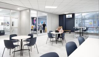 A modern, bright community space features tables and chairs, a reception area, and children interacting, creating a welcoming atmosphere.