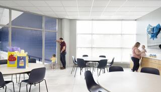 A bright room features a man with a child by the window, while a woman attends to another child and decorations on a counter.