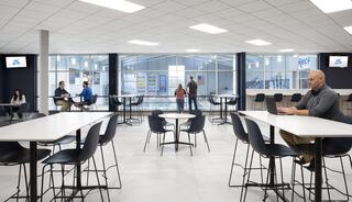 A modern, spacious area features simple tables and chairs, with people engaged in conversations and working, large windows allow natural light.